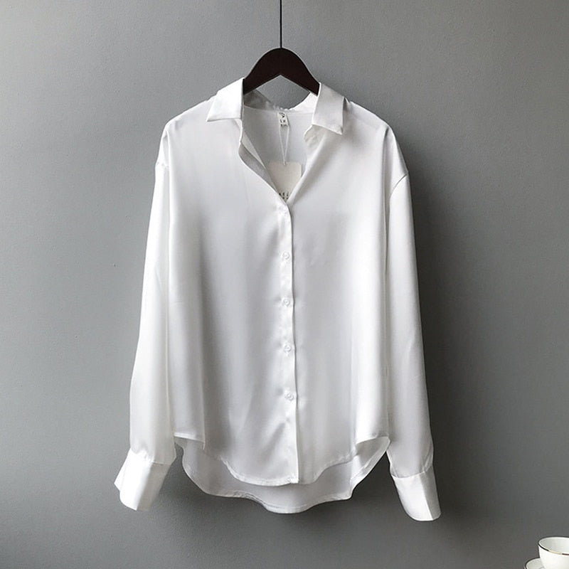 Une blouse élégante de couleur blanche suspendue à un cintre, avec un col classique et des manches longues retroussées, parfaite pour un look raffiné et intemporel.