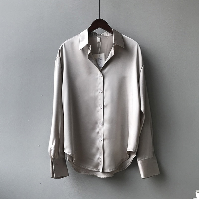 Une blouse en satin beige avec un col classique et des manches longues, suspendue à un cintre, offrant une finition élégante et fluide.