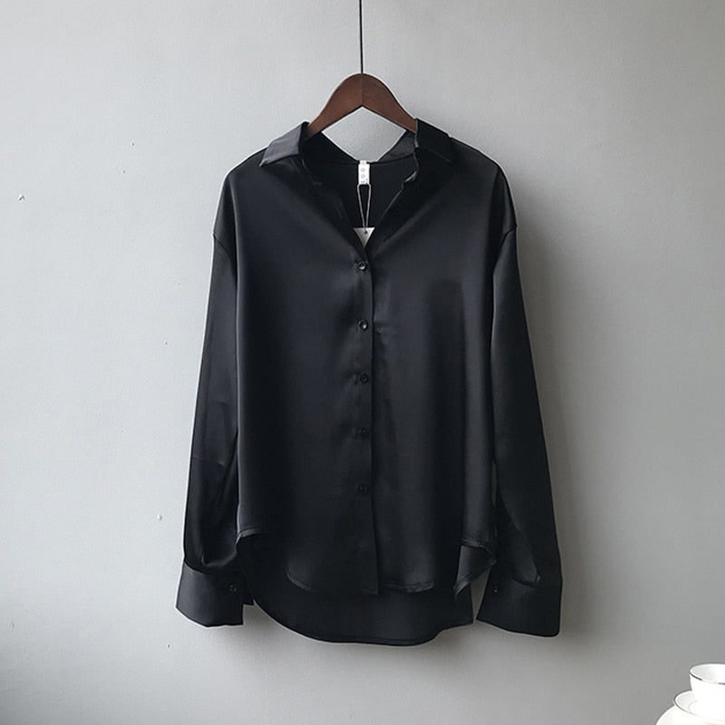 Une blouse en satin noir avec un col classique et des manches longues, suspendue à un cintre, montrant sa finition lisse et élégante.