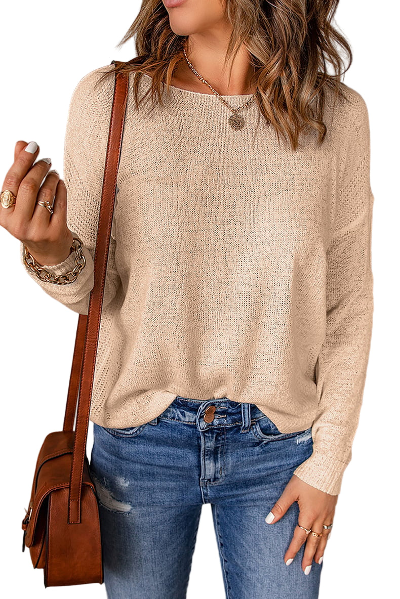 Une femme porte un pull décontracté à col rond beige, associé à un jean bleu clair légèrement déchiré. Elle tient un sac à bandoulière marron et porte plusieurs bagues, ainsi qu’un pendentif discret. Son regard est dirigé vers la droite.