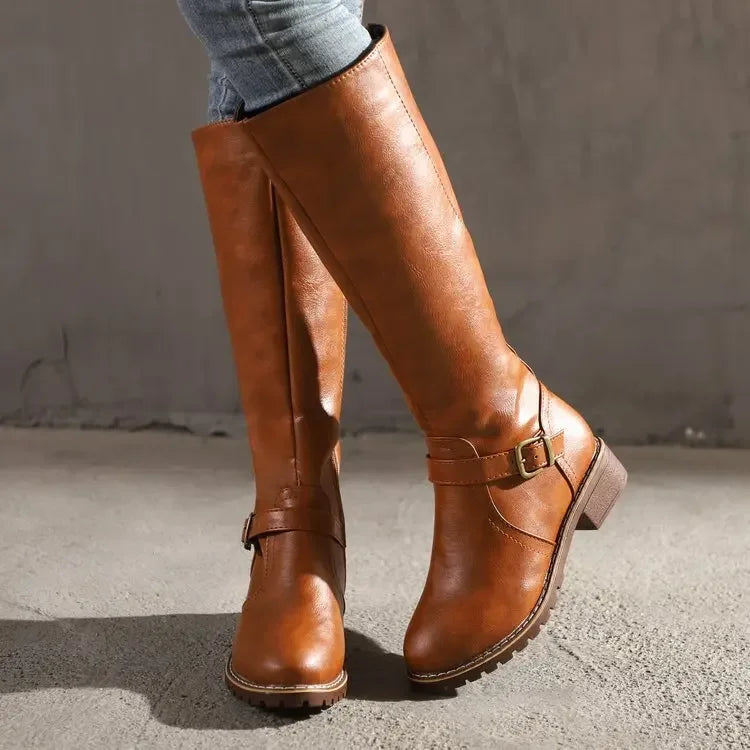 Paire de bottes hautes en cuir synthétique marron, dotées d’une fermeture éclair latérale et d’une bride avec boucle dorée à la cheville. Leur semelle crantée et leur talon bas assurent confort et stabilité. Ces bottes au style intemporel offrent une allure élégante et décontractée, parfaites pour l’automne et l’hiver, à porter avec un jean ou une jupe.