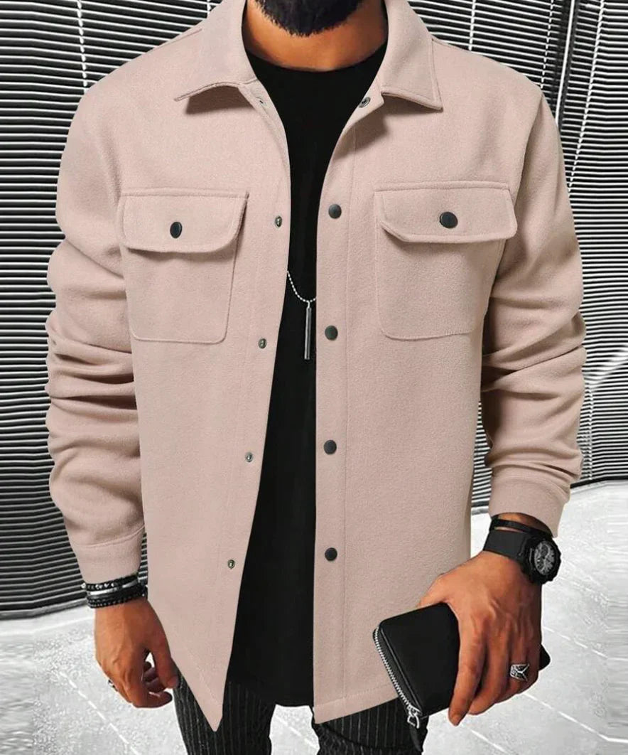 Homme portant une chemise beige à deux poches sur la poitrine, un t-shirt noir en dessous, une montre et un portefeuille en cuir, posé dans un environnement moderne.