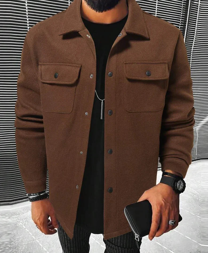 Homme portant une chemise marron à boutons noirs avec des poches sur la poitrine, associée à un t-shirt noir en dessous. Il porte des accessoires tels qu’une montre et des bracelets. Le fond épuré avec des lignes grises modernes contraste avec la simplicité et l’élégance de la chemise. Ce look combine confort et style décontracté, parfait pour une tenue raffinée et moderne.
