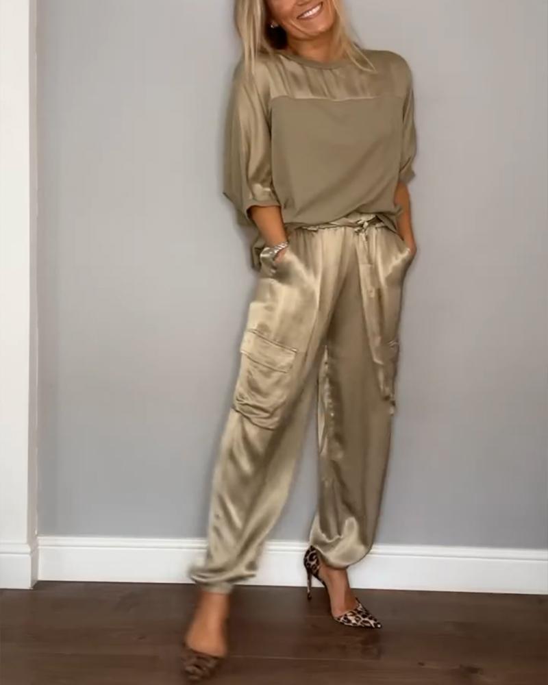 Une femme souriante portant un ensemble chic avec un haut beige fluide et un pantalon satiné beige avec des poches latérales. Elle porte des talons à motif léopard et se tient avec les mains dans les poches.