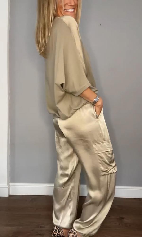 Une femme souriante portant un haut fluide beige et un pantalon satiné beige avec des poches latérales. Elle a un look décontracté, les mains dans les poches, et porte des talons à imprimé léopard.