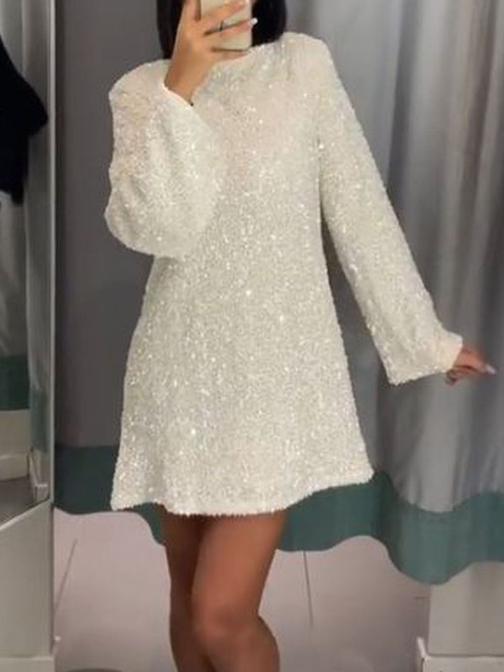 Une femme portant une robe blanche à sequins, avec des manches longues et un léger évasement au bas. Elle prend un selfie dans un miroir, montrant la texture brillante de la robe et sa coupe élégante.