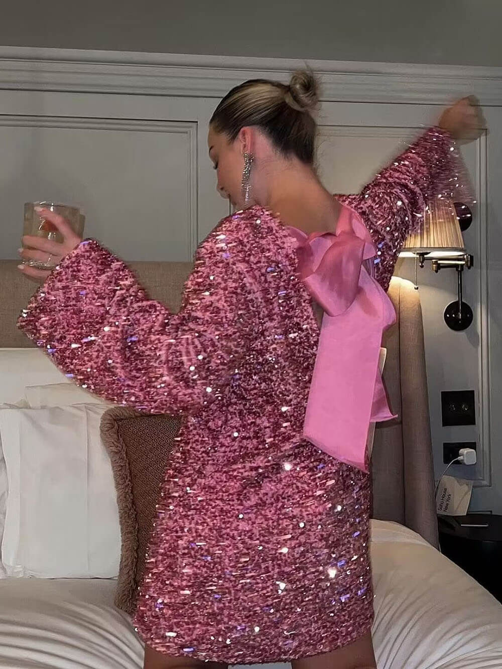 Une femme dans une robe rose à sequins, avec un large nœud en satin rose dans le dos. Elle tient un verre dans une main et se tourne, les bras étendus, posant sur un lit. Le fond montre une chambre élégante, avec des coussins et des lampes.