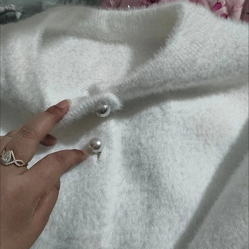 Gros plan d'une main ajustant les boutons en perle d'un cardigan blanc en tricot doux. Le tissu duveteux et les détails des boutons ressortent clairement sur l'image.