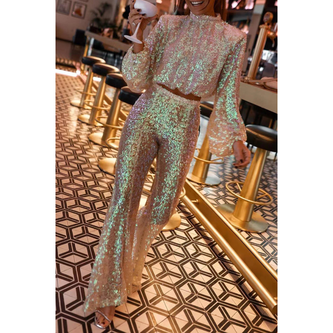 Femme portant un ensemble de soirée scintillant composé d’un haut à manches longues et d’un pantalon évasé assorti, tous deux recouverts de sequins irisés aux reflets dorés et verts. Le haut présente un col montant et une coupe légèrement bouffante, tandis que le pantalon taille haute allonge la silhouette. La tenue reflète la lumière avec élégance, créant un effet glamour idéal pour les fêtes ou les soirées modernes.