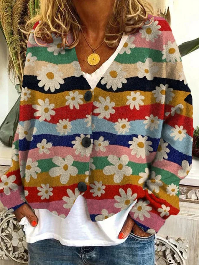 Femme portant un cardigan à motifs floraux multicolores avec de grandes marguerites blanches sur des bandes ondulées rouges, vertes, bleues et dorées. Le vêtement est ample, à manches longues, avec de gros boutons noirs sur le devant. Elle porte un t-shirt blanc en dessous et un jean décontracté, créant un style bohème et vintage.