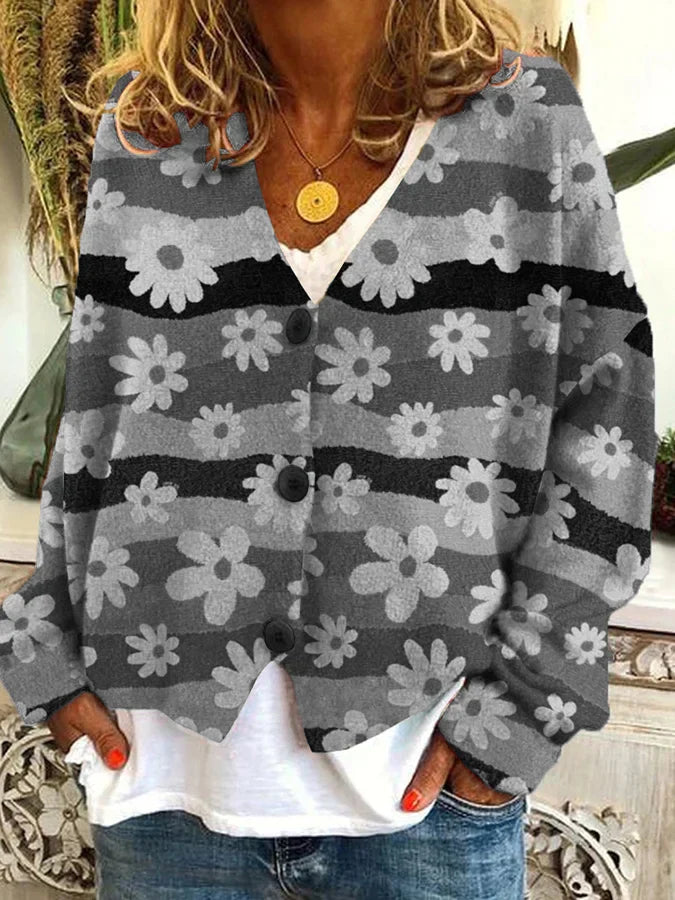 Femme portant un cardigan à boutons au style vintage, orné de larges rayures horizontales dans différentes nuances de gris et de noir, décoré de motifs de fleurs blanches. Le vêtement est porté avec un t-shirt blanc décontracté et un jean bleu, créant un look à la fois rétro et moderne. Le tissu du cardigan semble doux et confortable, parfait pour une tenue automnale ou hivernale.