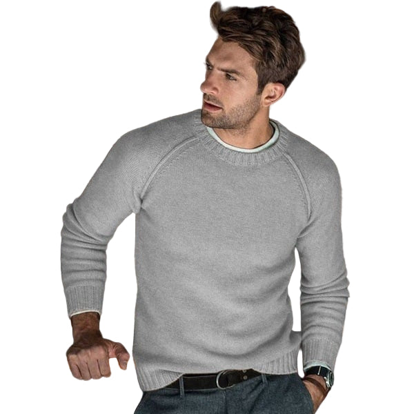 Un homme portant un pull en tricot gris clair avec un col rond. Il a un look moderne et élégant, avec des cheveux courts et une posture détendue, vêtu d'un pantalon gris et d'une ceinture noire, posé dans un environnement intérieur chaleureux.