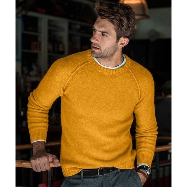 Un homme portant un pull tricoté jaune avec un col rond. Il se tient debout, appuyé sur une barre dans un environnement intérieur, portant des pantalons gris et une ceinture noire. Il regarde sur le côté avec une expression pensive.