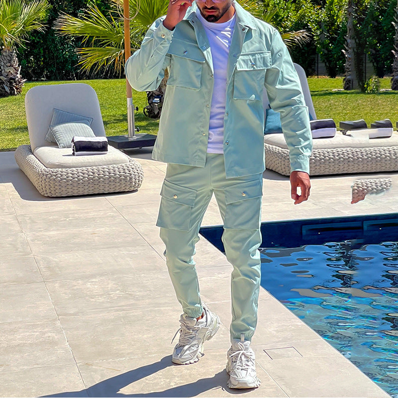 Un homme porte un ensemble cargo bleu clair, composé d'une chemise à boutons et d'un pantalon cargo assorti. Il marche près d'une piscine, avec des chaises longues en arrière-plan et un environnement extérieur moderne. Il porte des baskets blanches et un t-shirt blanc sous la chemise.