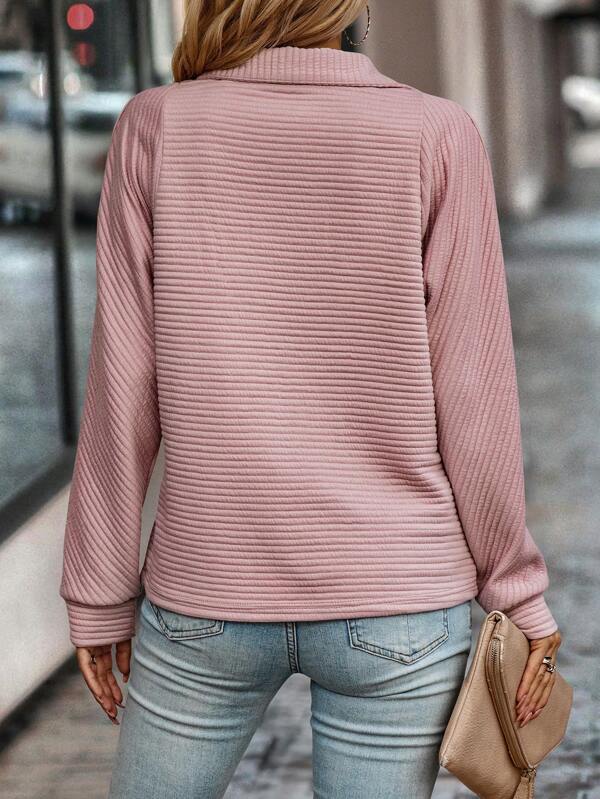 Vue de dos d'une femme portant un pull rose pâle à manches longues et à mailles côtelées horizontales. Ce pull, à la coupe droite et au col montant, est associé à un jean bleu clair et une pochette beige. Le style est féminin, simple et élégant, idéal pour un look moderne et décontracté.