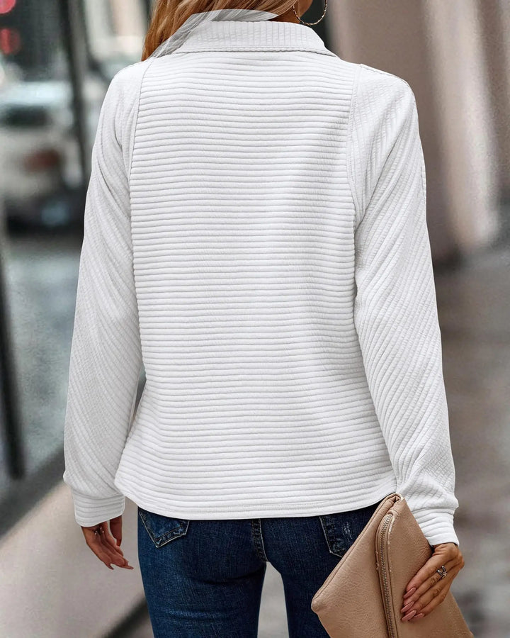 Vue arrière d’une femme portant un pull blanc à manches longues avec une texture côtelée horizontale. Le vêtement présente une coupe droite et un col montant, associé à un jean bleu et une pochette beige. Le style est simple, moderne et élégant, idéal pour un look chic au quotidien.