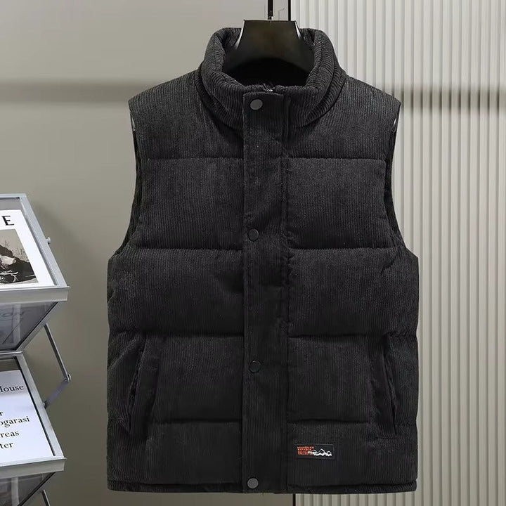 Gilet matelassé pour homme en velours côtelé de couleur noire, présentant une fermeture à boutons-pression et deux poches latérales discrètes. Le tissu texturé en velours offre une allure chic et moderne tout en assurant confort et chaleur pour la saison froide. Ce vêtement sans manches est parfait pour un style décontracté et élégant à la fois.