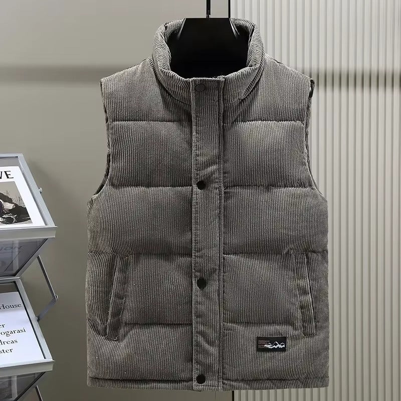 Gilet matelassé pour homme en velours côtelé gris, avec une fermeture à boutons-pression et deux poches latérales. Le col montant et la texture côtelée apportent une touche de style moderne et raffinée. Ce vêtement sans manches offre un équilibre parfait entre confort, chaleur et élégance, idéal pour la saison automne-hiver.