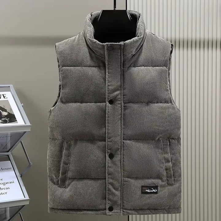 Gilet matelassé pour homme en velours côtelé gris, avec une fermeture à boutons-pression et deux poches latérales. Le col montant et la texture côtelée apportent une touche de style moderne et raffinée. Ce vêtement sans manches offre un équilibre parfait entre confort, chaleur et élégance, idéal pour la saison automne-hiver.