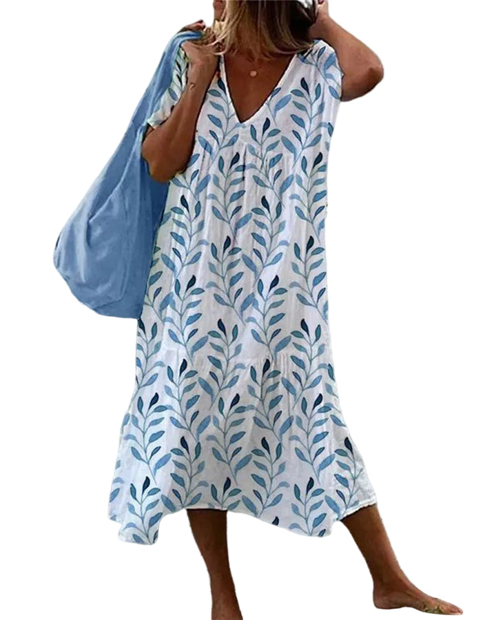 Robe col V pour femme Adèle, tendance, blanche à imprimé feuilles bleues. Coupe midi fluide et ample avec manches courtes et encolure en V, idéale pour un look estival léger, portée avec un grand sac bleu.