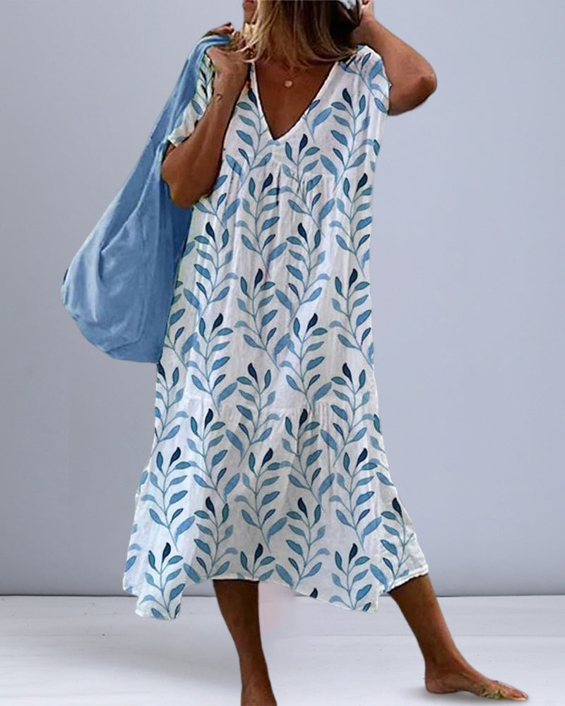 Robe col V pour femme Adèle, tendance, blanche avec imprimé feuillage bleu. Robe midi ample et fluide à manches courtes et décolleté en V, portée avec un grand sac bleu pour un style estival décontracté.