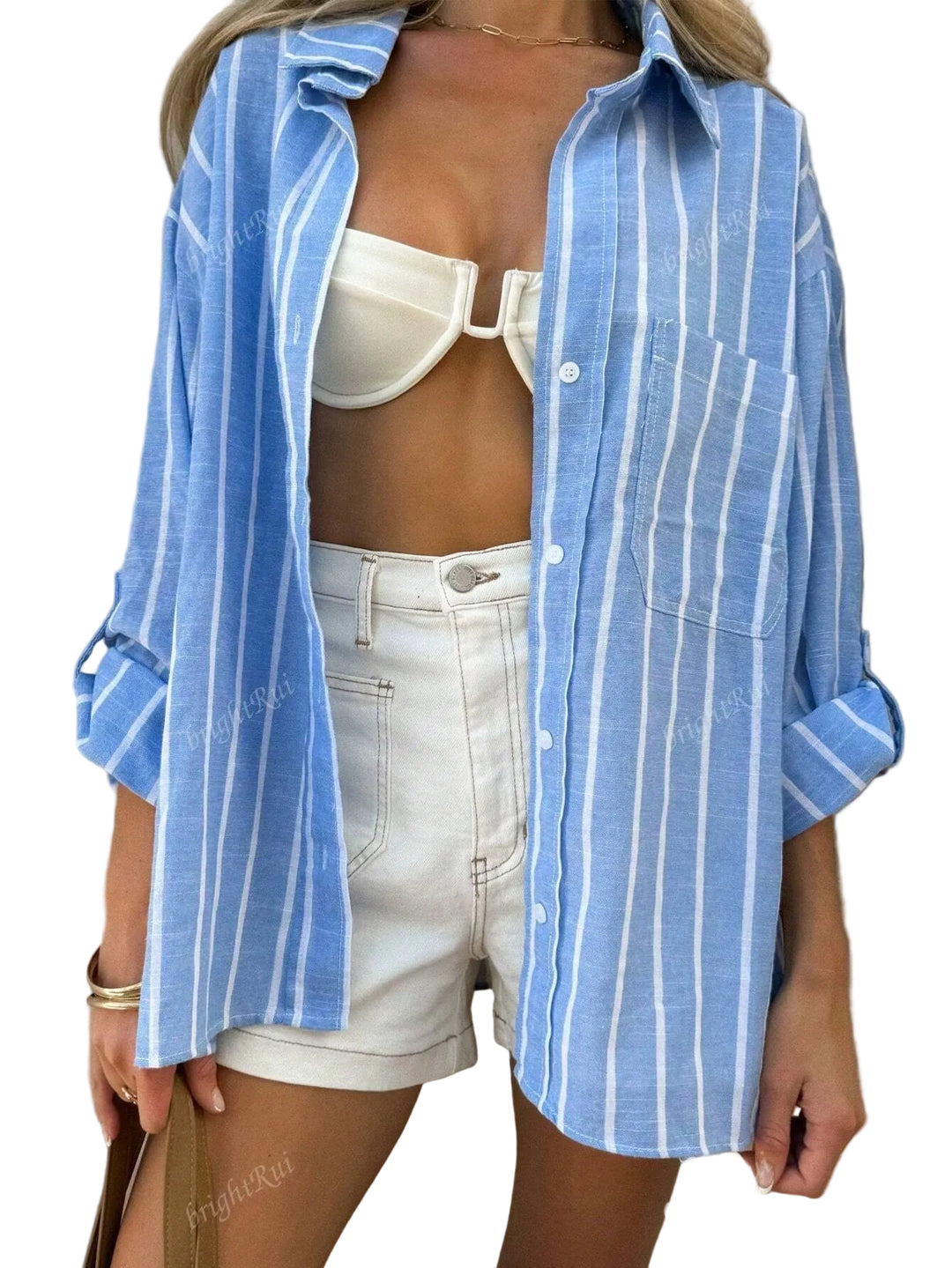 Le modèle porte une chemise oversize à rayures bleues et blanches, ouverte sur un bikini blanc. Elle associe la chemise à un short en jean blanc, créant un look estival et décontracté. Des bracelets dorés et un sac à main ajoutent une touche chic à cet ensemble moderne et stylé.