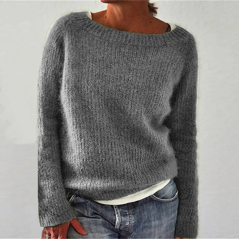 Femme portant un pull gris en maille douce à manches longues, avec une coupe décontractée et une encolure large, superposé à un t-shirt clair et associé à un jean, photographiée sur un fond neutre.