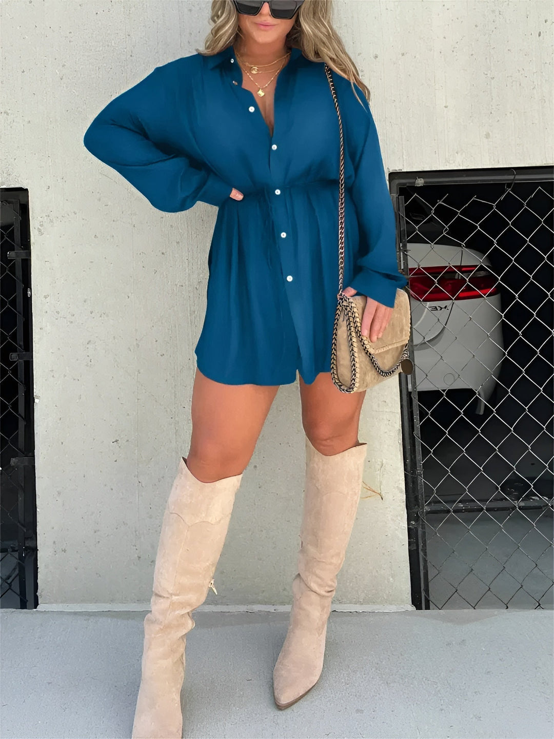 Chemise à manches longues pour femme, modèle tendance. Cette chemise fluide de couleur bleu canard, dotée de boutons et d'une ceinture à nouer à la taille, crée une silhouette élégante et moderne. Elle est portée avec des bottes hautes beiges et un sac à bandoulière, parfait pour un look chic et décontracté.