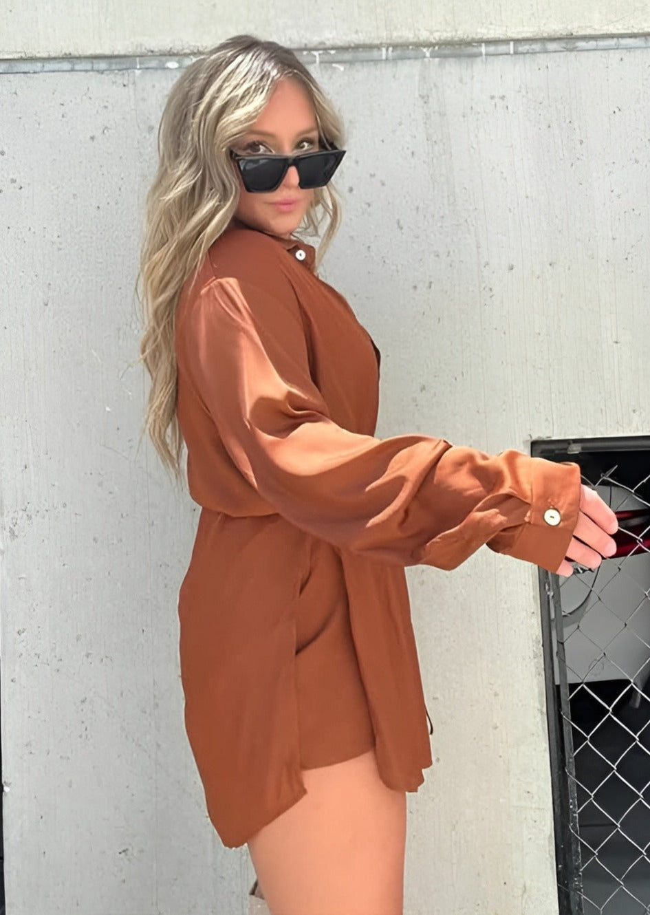 Chemise à manches longues pour femme, modèle tendance. Cette chemise fluide de couleur marron terracotta, avec des boutons et une ceinture à nouer à la taille, offre un look élégant et décontracté. Elle est portée avec des lunettes de soleil et un sac à bandoulière, parfait pour un style moderne et chic.