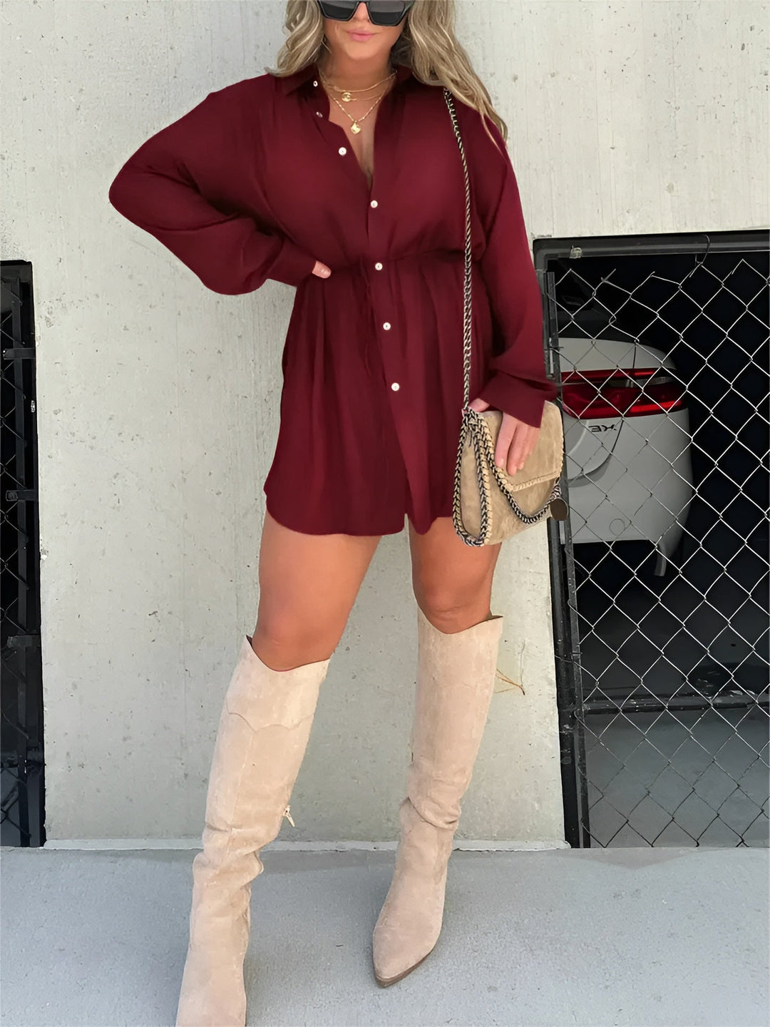 Chemise à manches longues pour femme, modèle tendance. Cette chemise fluide de couleur bordeaux, dotée de boutons et d'une ceinture à nouer à la taille, crée une silhouette chic et moderne. Elle est portée avec des bottes hautes beiges et un sac à bandoulière, parfait pour un look élégant et décontracté.
