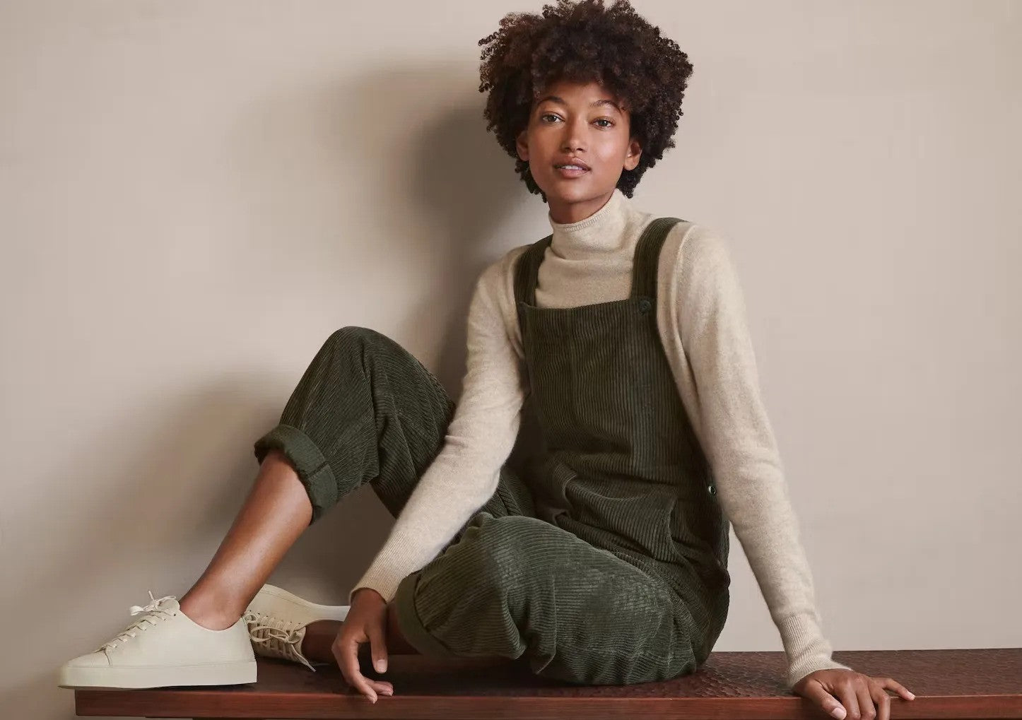 Femme portant une salopette en velours côtelé vert olive avec un pull beige à col roulé, assise sur une table en bois et portant des baskets blanches.