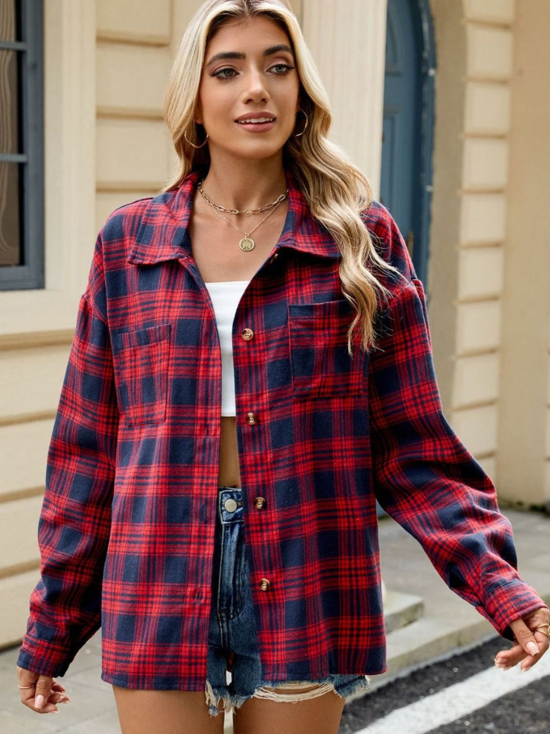 Femme portant une chemise longue à carreaux rouge et bleu, un débardeur blanc et un short en jean déchiré, posant dans un environnement moderne avec un fond de mur beige.