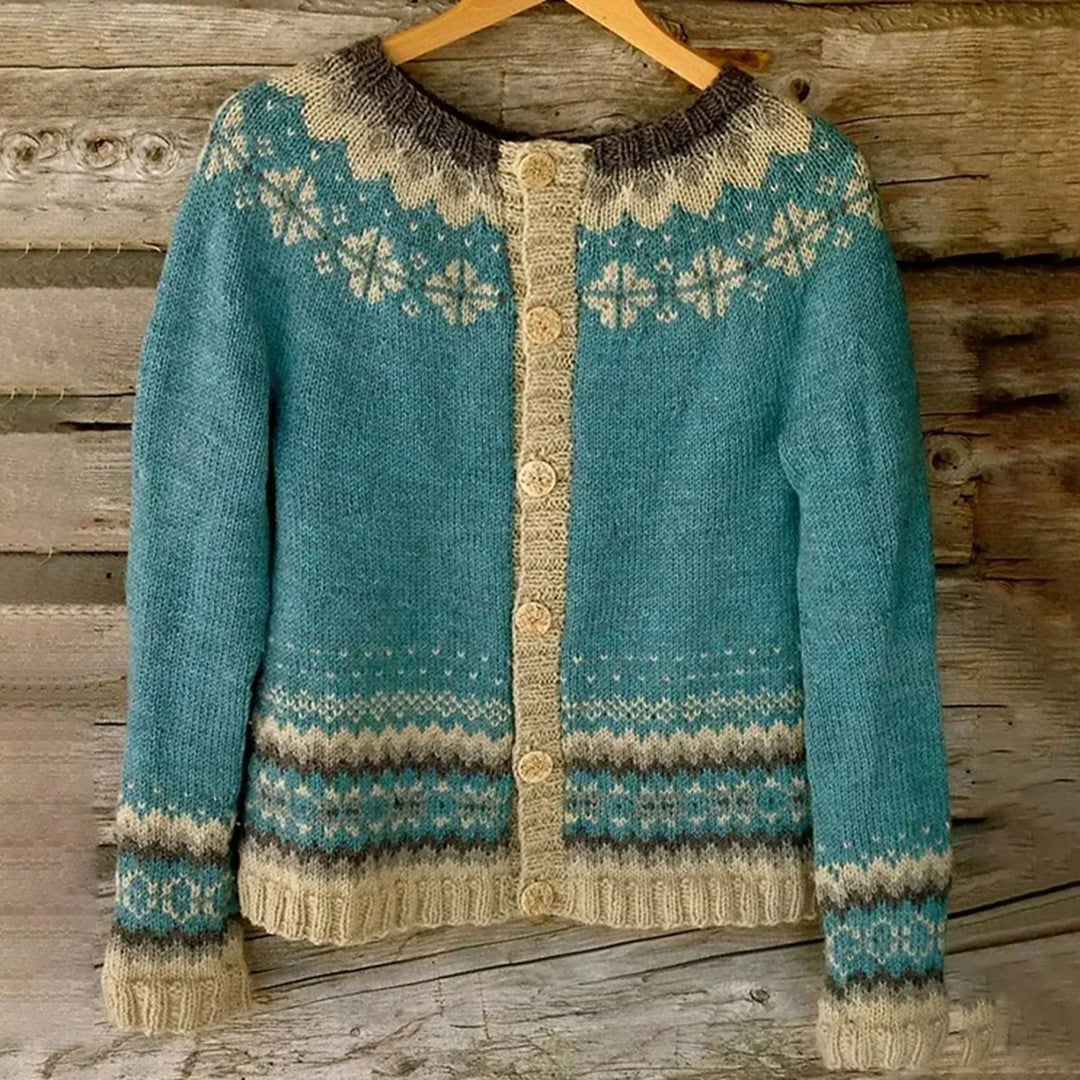 Cardigan en maille turquoise suspendu à un cintre, orné de motifs nordiques beige et gris sur le col, le bas et les poignets, avec une rangée de boutons tricotés assortis, photographié devant un fond en bois rustique.