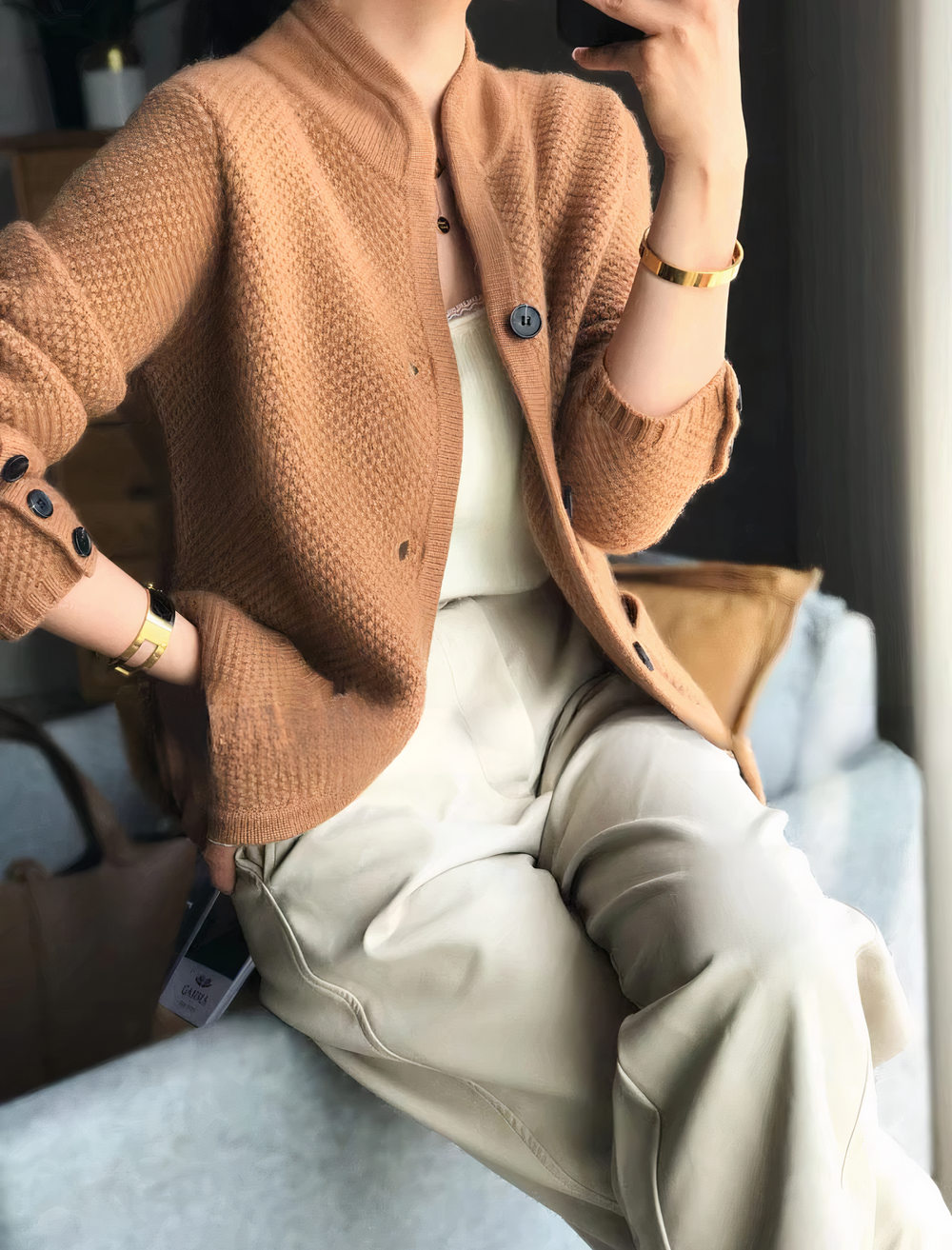 Femme assise portant un cardigan moderne de couleur camel, à maille texturée et boutons contrastés, associé à un haut léger et un pantalon beige pour un look élégant et décontracté.