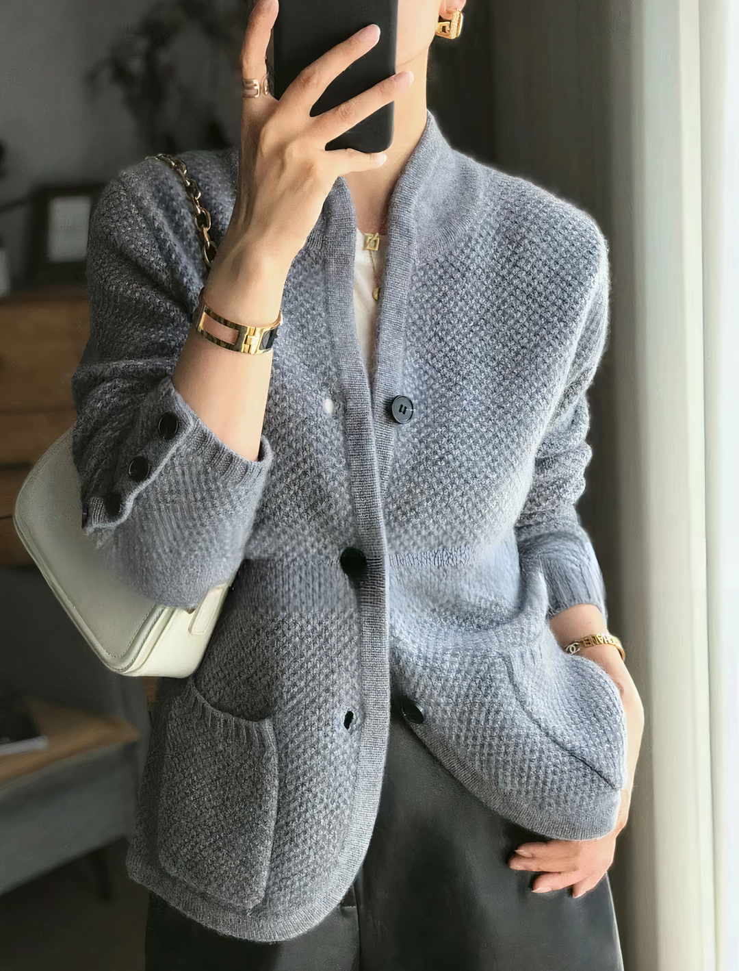 Femme portant un cardigan moderne gris à maille texturée, doté de grands boutons contrastés et de poches avant, assorti à un sac élégant et des accessoires dorés pour un style chic et minimaliste.