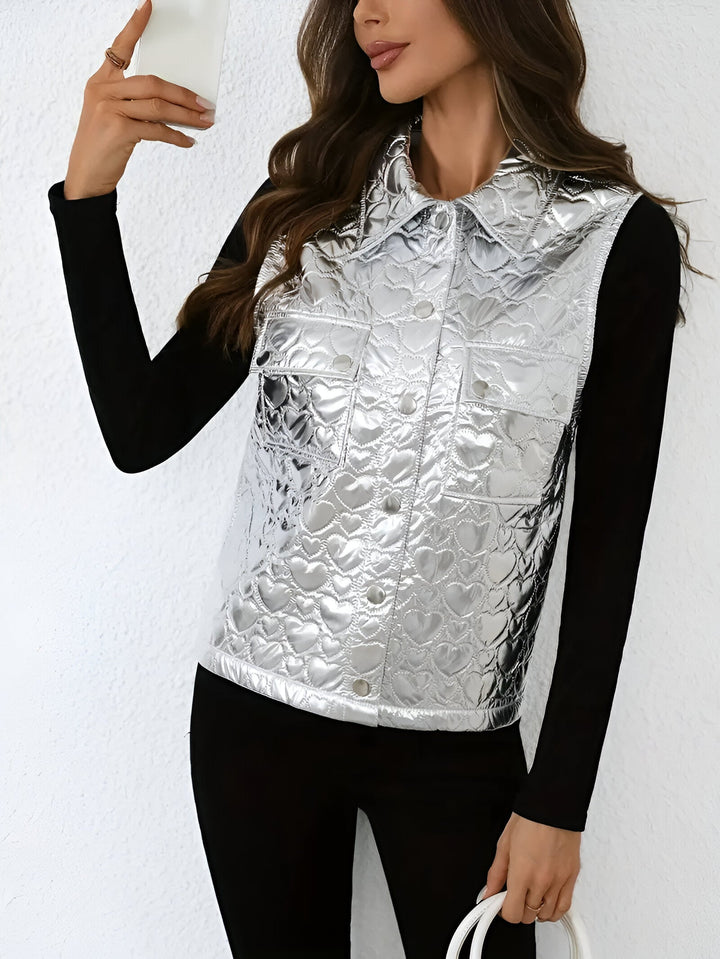 Femme prenant un selfie, vêtue d’un gilet sans manches argenté au fini métallisé avec un motif matelassé en forme de cœur. Elle le porte sur un haut noir ajusté et tient un petit sac blanc. Son look moderne et élégant met en valeur un style audacieux et tendance.