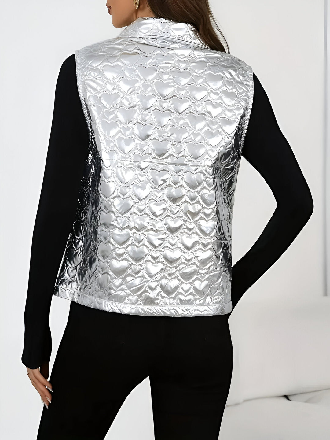 Vue de dos d’une femme portant un gilet sans manches argenté à effet métallisé avec un motif matelassé en forme de cœur. Le vêtement brille sous la lumière et se marie avec un haut noir ajusté et un pantalon assorti, créant un style moderne et élégant.