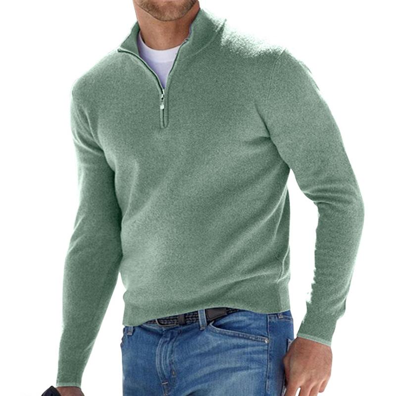 Pull pour homme vert sauge avec col montant et fermeture zippée courte, coupe ajustée à manches longues, porté avec un jean bleu sur fond blanc.