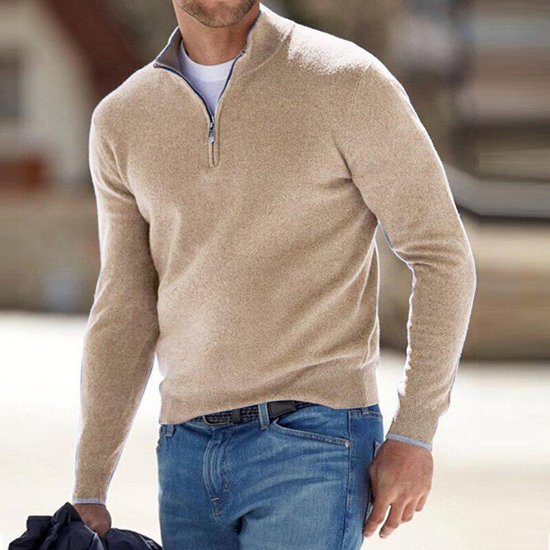 Pull pour homme beige avec col montant et fermeture zippée courte, coupe ajustée à manches longues, porté avec un jean bleu dans un décor extérieur.