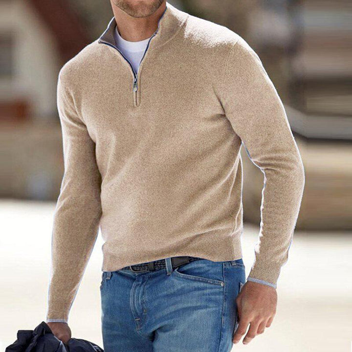 Pull pour homme beige avec col montant et fermeture zippée courte, coupe ajustée à manches longues, porté avec un jean bleu dans un décor extérieur.