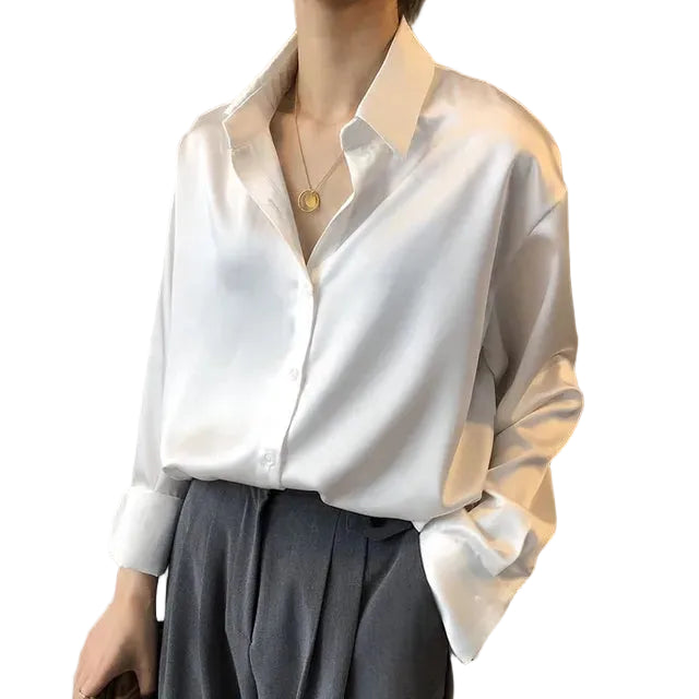 Chemisier en satin blanc pour femme, avec un col classique et des boutons sur le devant. Le vêtement a une coupe ample, les manches sont retroussées aux poignets, et il est associé à un pantalon gris élégant. Parfait pour un look décontracté et chic, idéal pour le bureau ou une sortie en ville.