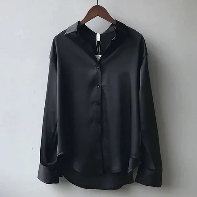 Chemisier en satin noir pour femme, avec un col classique et des boutons sur le devant. Le vêtement présente une coupe ample avec des manches longues et des poignets légèrement retroussés. Idéal pour un style élégant et raffiné, parfait pour une tenue professionnelle ou une soirée chic.