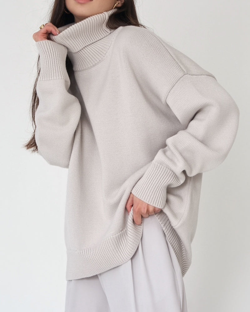 Femme portant un pull à col roulé ample de couleur gris clair, au tricot épais et doux. Le vêtement présente une coupe oversize avec des manches longues et des finitions côtelées au col, aux poignets et à la base. Le style moderne et minimaliste crée une allure à la fois élégante et décontractée, idéale pour les journées d’automne ou d’hiver. Le fond clair met en valeur la texture du tissu et la douceur de la teinte neutre.