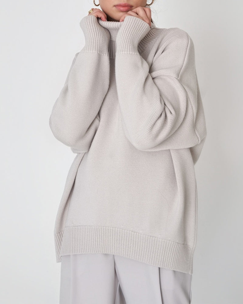 Femme portant un pull à col roulé ample de couleur gris clair, au tissu tricoté doux et à la coupe décontractée. Les manches longues et légèrement tombantes ajoutent une touche confortable et élégante. Le style minimaliste et moderne convient parfaitement aux journées fraîches, offrant une allure à la fois chic et décontractée. L’arrière-plan blanc met en valeur la texture du pull et la douceur du ton neutre.