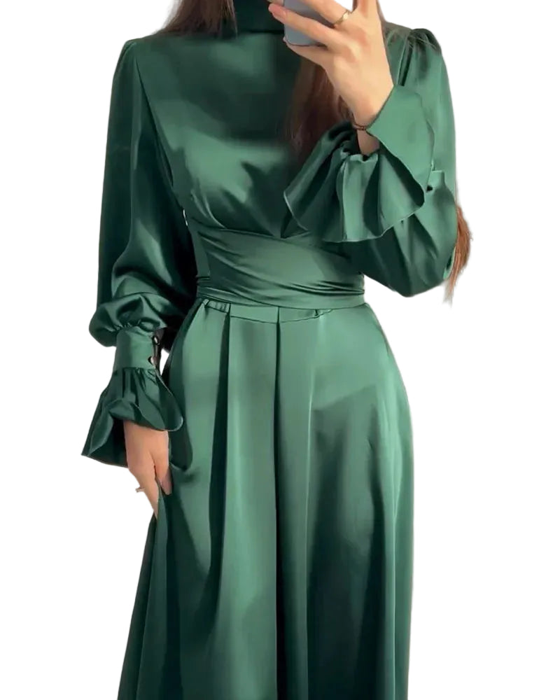 Robe longue pour femme Aveline vert foncé, col montant, taille cintrée et jupe plissée, manches longues avec volants aux poignets, portée en intérieur.