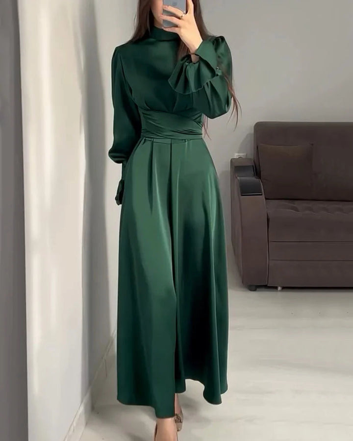 Robe longue pour femme Aveline vert foncé, manches longues et col montant, taille cintrée avec plis, portée en intérieur devant un canapé.