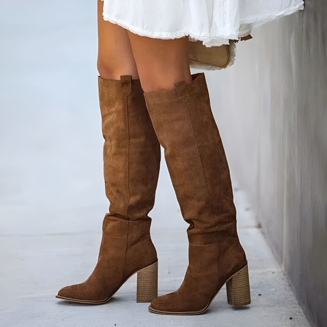 Gros plan sur des bottes hautes en daim marron à talons épais portées avec une robe blanche légère, photographiées le long d’un mur extérieur sur un sol clair.