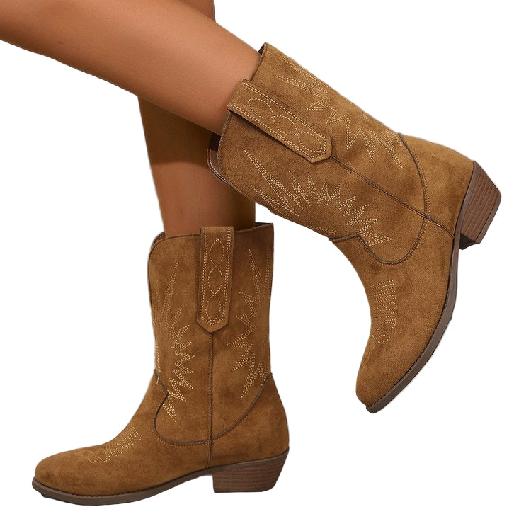 Bottes style cowboy en daim marron avec broderies décoratives, portées par une femme posant sur un sol en bois vieilli devant un fond rustique en planches.