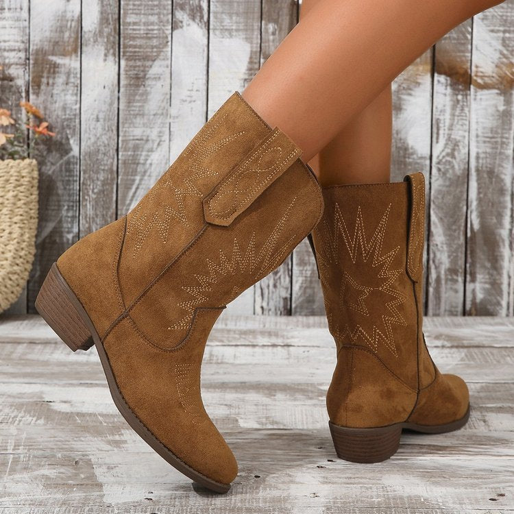 Bottes style cowboy en daim marron avec broderies décoratives sur la tige et l’arrière, portées par une femme posant sur un sol en bois vieilli devant un fond en planches rustiques.