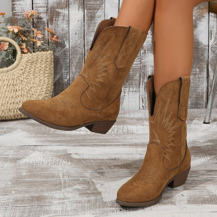 Bottes style cowboy en daim marron avec broderies décoratives, portées par une femme assise sur une chaise, la jambe relevée, sur un sol en bois patiné devant un fond en planches rustiques.
